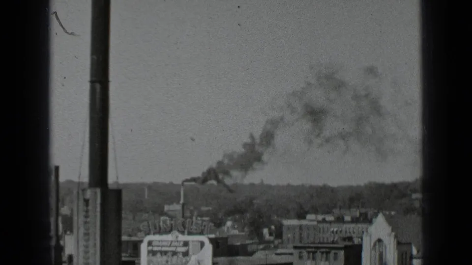 Black-and-white 8mm footage frames a New York urban skyline with a chimney plume crossing above low buildings.
