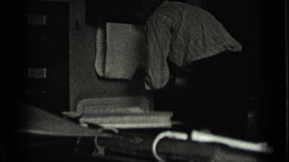 Black-and-white 8mm frame showing a dim interior or porch area with furniture, a draped cloth, and strong shadow.