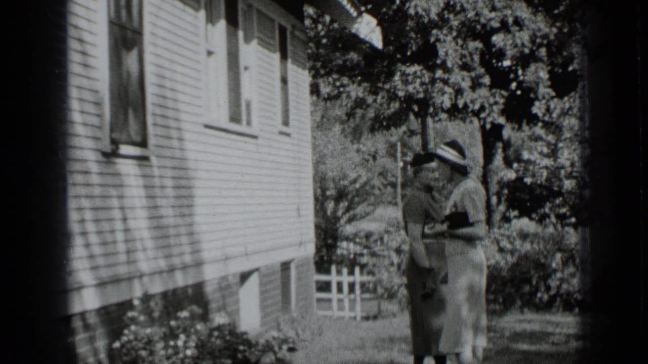 Two adult women stand or walk beside a clapboard house, shrubs, and trees.