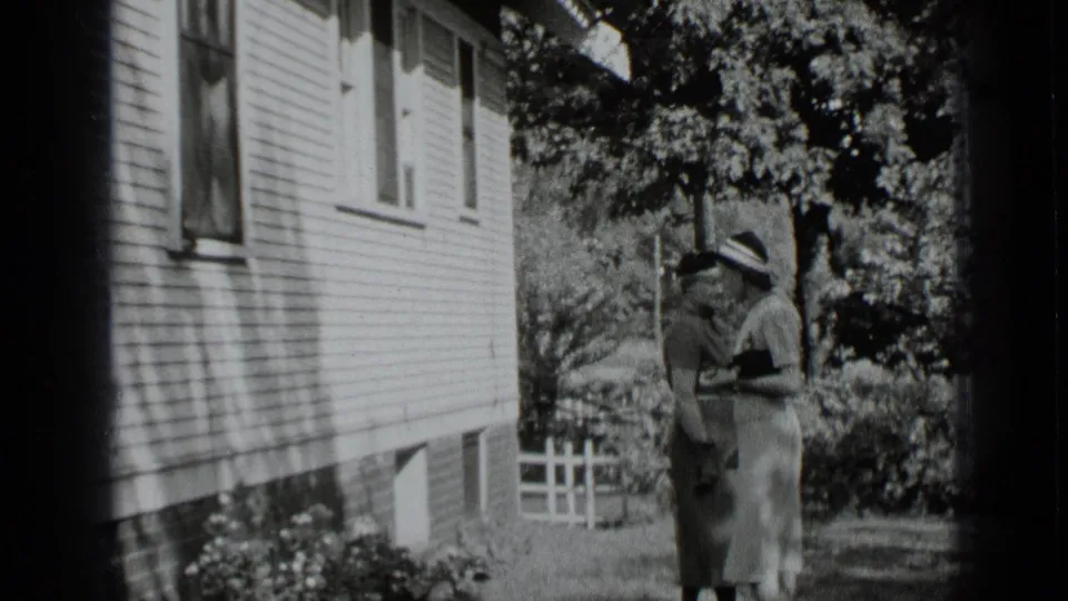 1939 New York home-movie frame of two adult women in dresses and hats near a clapboard house and garden shrubs.