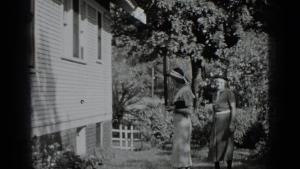 1939 New York black-and-white 8mm frame of two women strolling near a residential garden and picket fence.