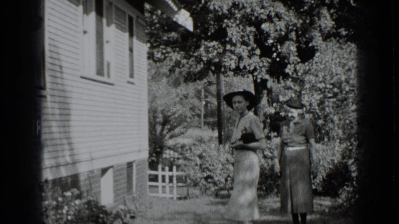 Two women in dresses and brimmed hats stand beside a clapboard house in a garden.