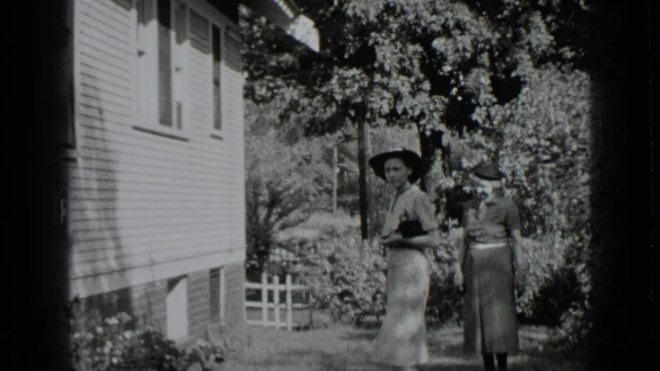 Two women in 1939 New York home-movie footage stand in a sunlit garden beside a clapboard house and picket fence.