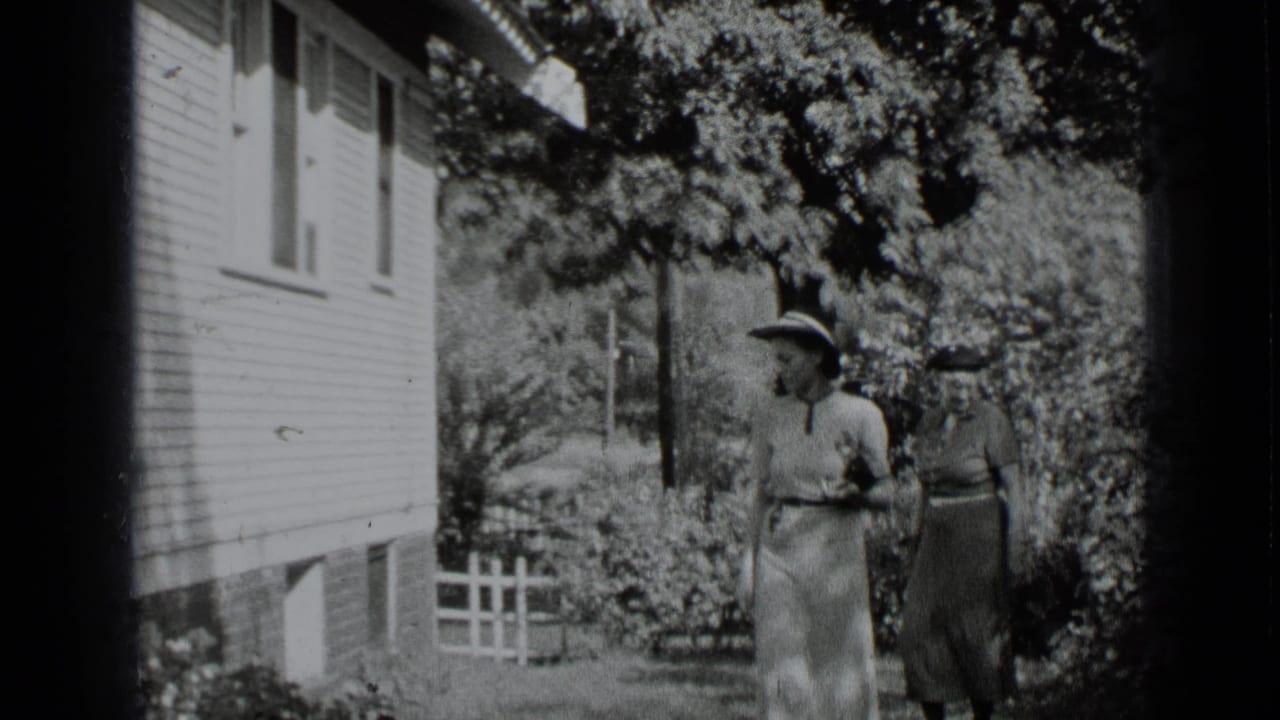 Two women walk or stand in a garden beside a light-colored house and fence.