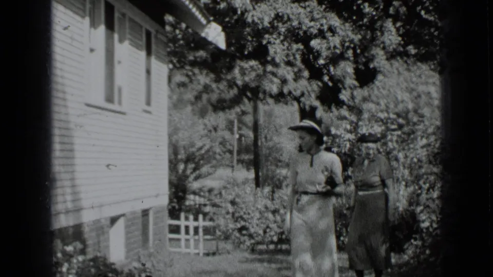Two women in brimmed hats appear in a 1939 New York garden scene beside a clapboard house and white picket fence.