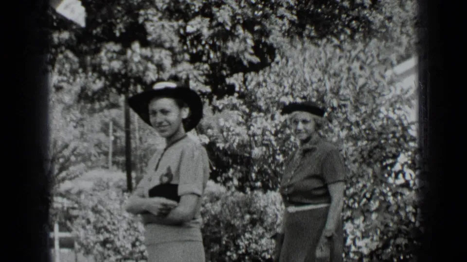 Closer 1939 New York home-movie frame of two smiling women in hats, one holding a small dark item, with shrubs behind them.