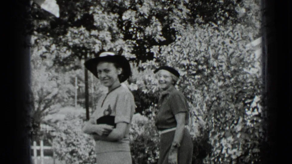 Two women in 1939 New York footage smile toward the camera, framed by dense shrubs and dappled sunlight.