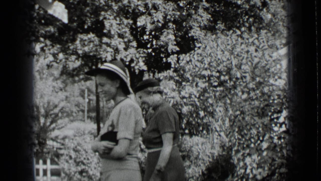 Two women in hats stand near shrubs in a garden.