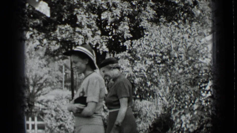 Two women in period hats and skirts pause beside shrubs in a 1939 New York home-movie garden scene.