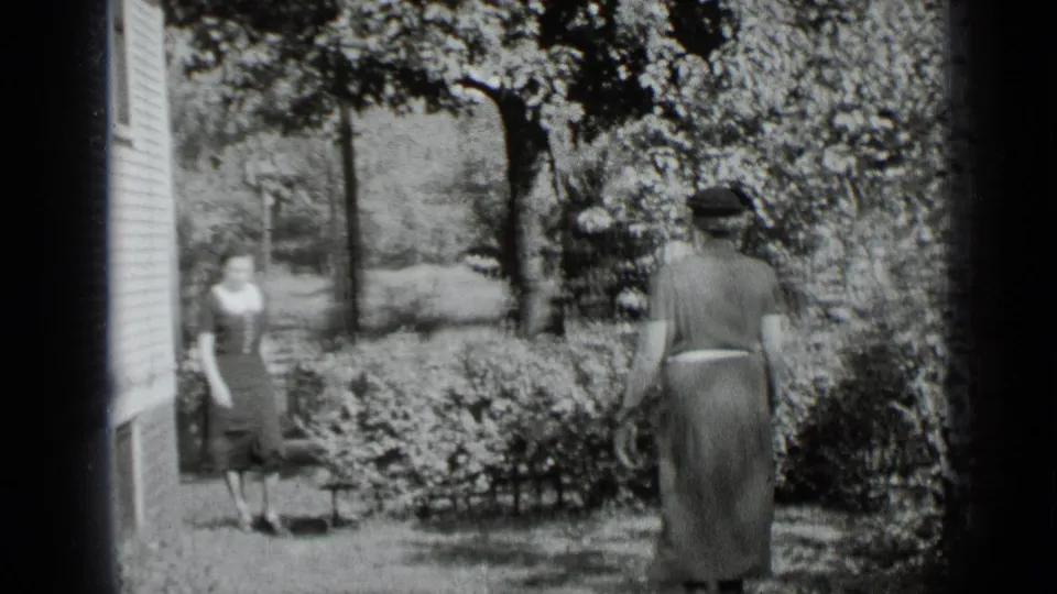 In a leafy New York yard, one woman walks toward another standing near the side of a house.
