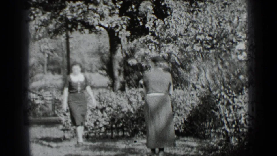 Two women in 1930s-style clothing face each other in a New York garden, with shrubs and trees behind them.