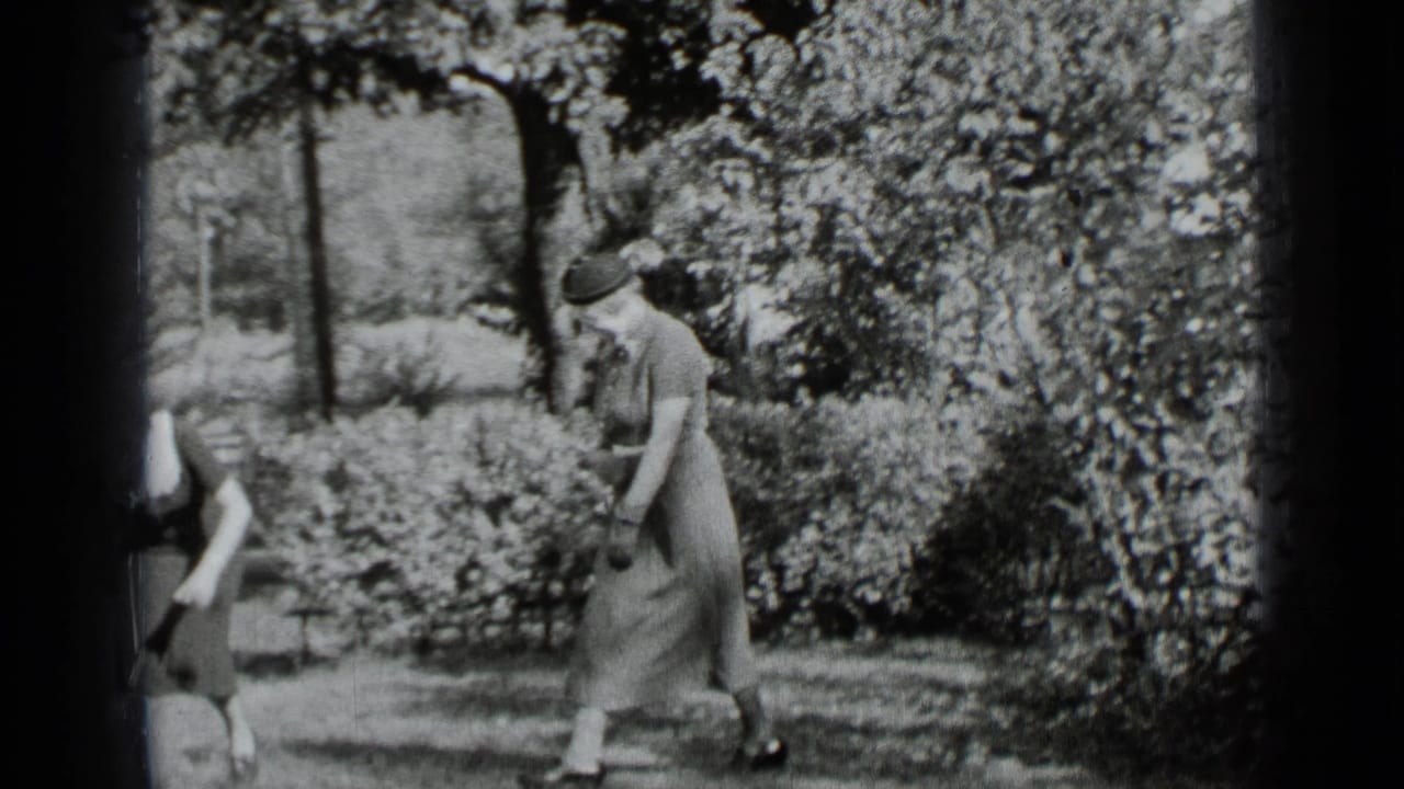 Woman in a long dress and hat walks across a sunny yard beside hedges and trees.