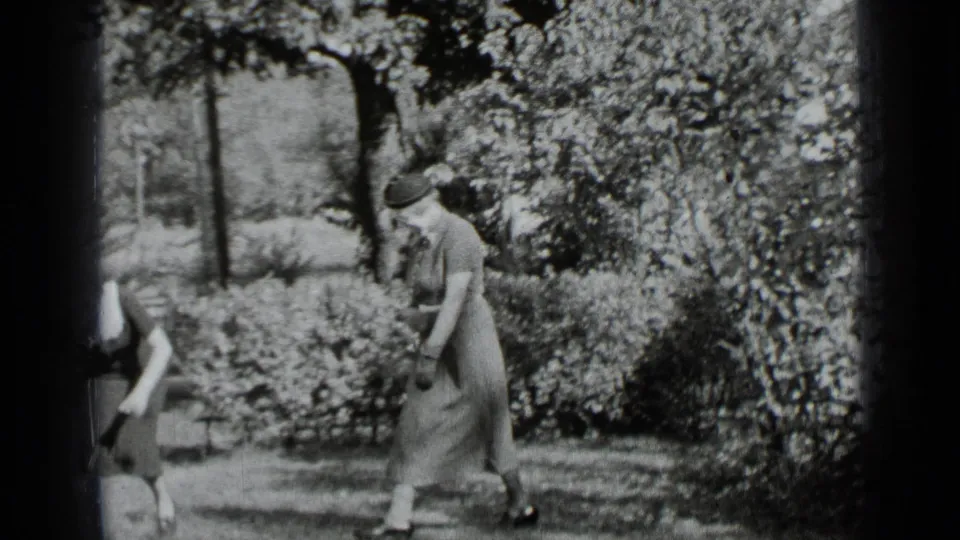 In 1939 New York home-movie footage, a woman in a long dress and hat moves through a residential yard bordered by hedges and trees.