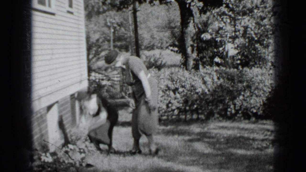 Two women stand near the side of a clapboard house, with one bending toward plants.