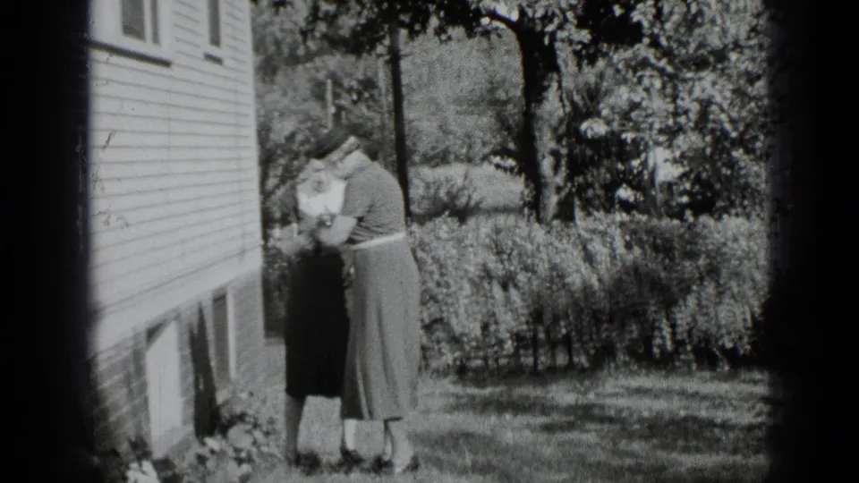 Two women in vintage dress stand near the clapboard house wall and garden edge in black-and-white 8mm footage from New York.