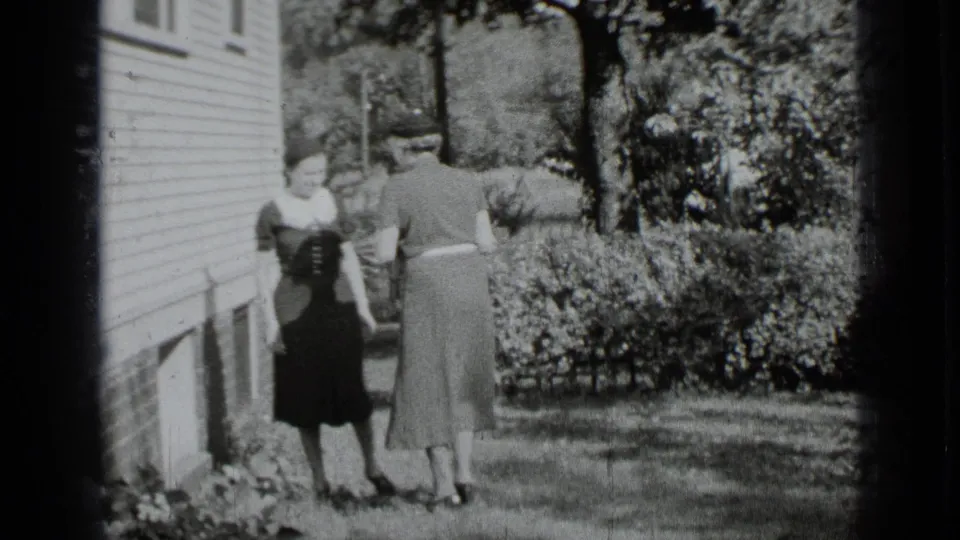 The two women face each other near hedges and the clapboard house exterior, recorded in 1939 New York home-movie footage.