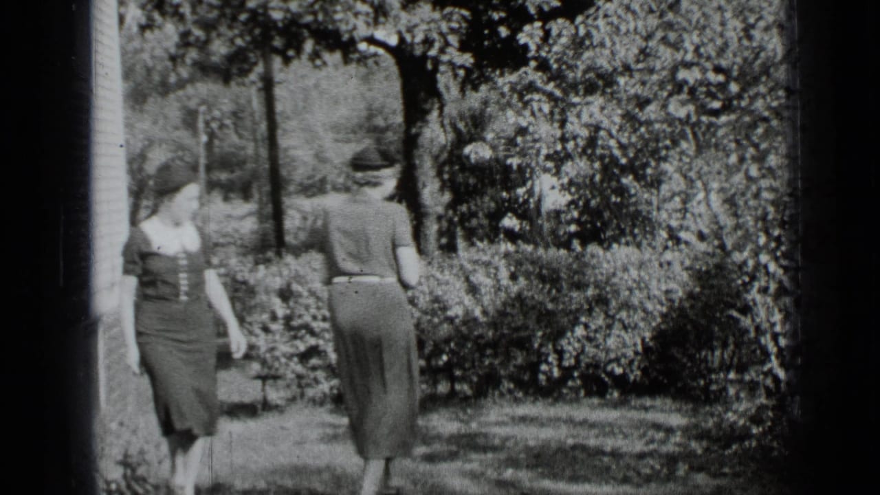 Two women in dresses and hats meet on a sunny garden lawn beside a house.
