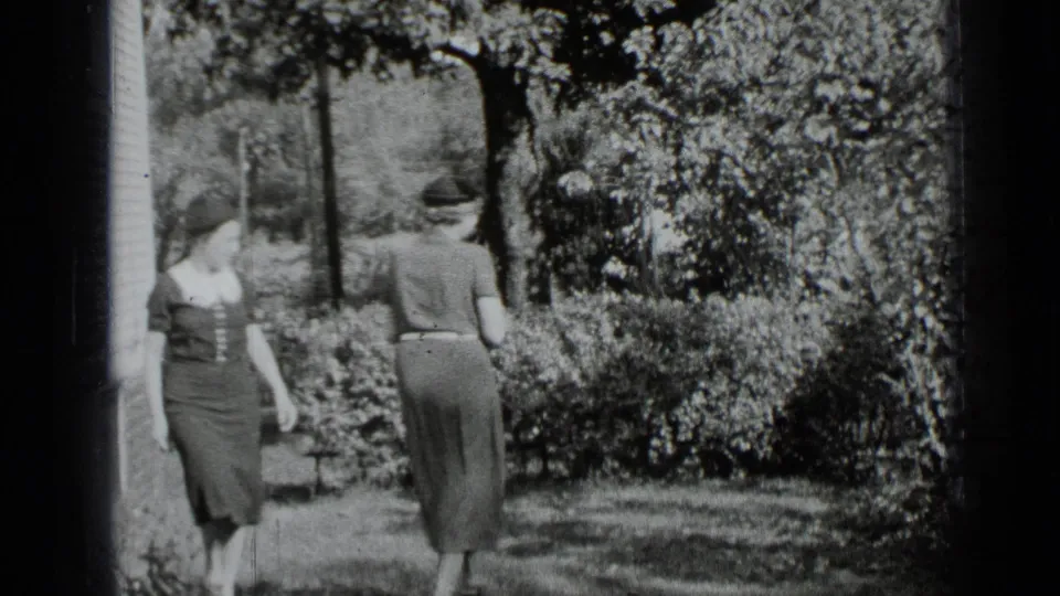 Two women in 1939 New York home-movie footage stand near a clapboard house and garden shrubs during an outdoor greeting.