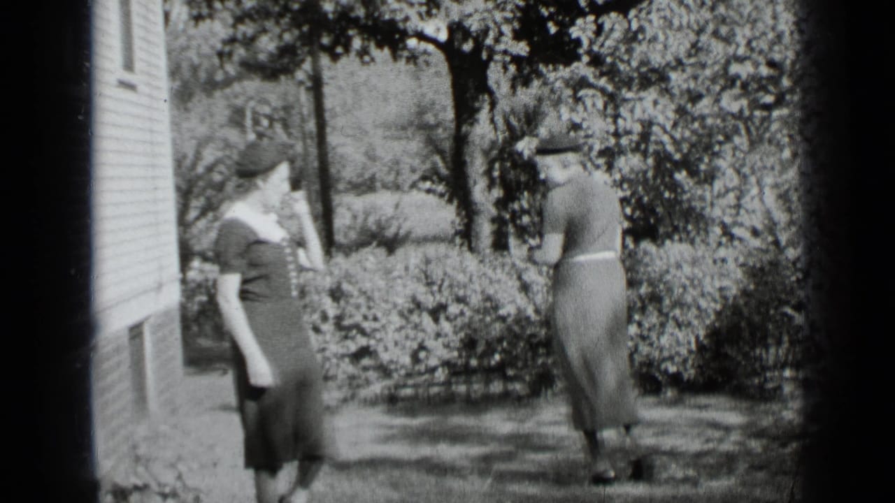 Two women stand near shrubs and a clapboard house in bright sun.