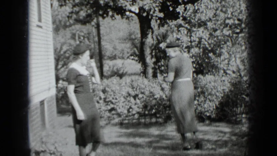 Two women in vintage dresses and hats stand outdoors near garden shrubs and a house, consistent with the 1939 New York metadata.