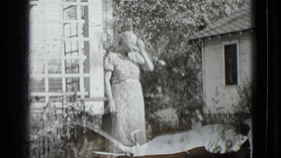 A woman in a light patterned dress stands outside near porch lattice, flowers, and a small outbuilding in 1939 New York home-movie footage.