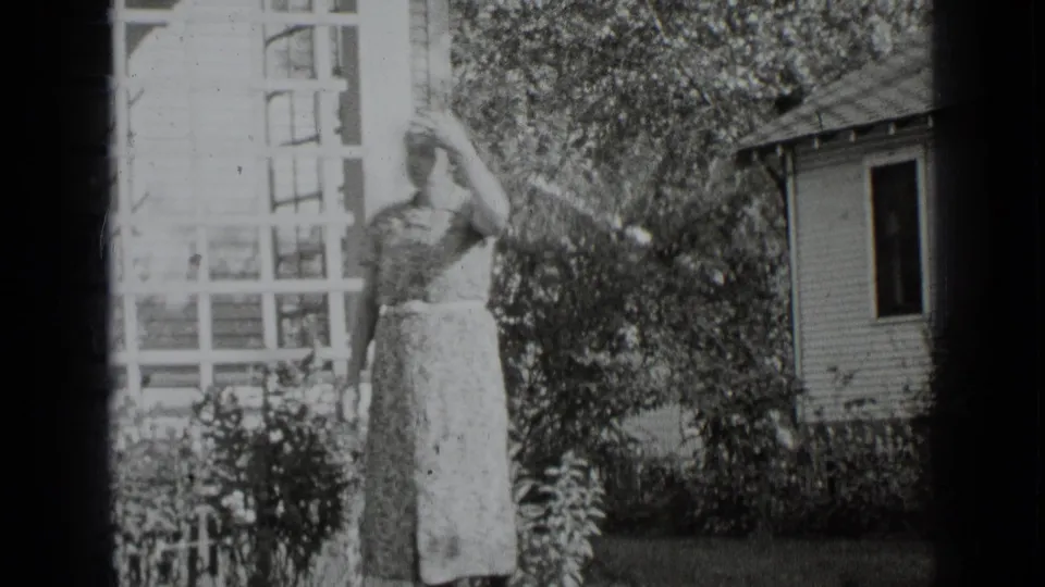 A woman in a long patterned dress stands in a sunny garden near porch lattice and clapboard siding, matching the supplied 1939 New York context.