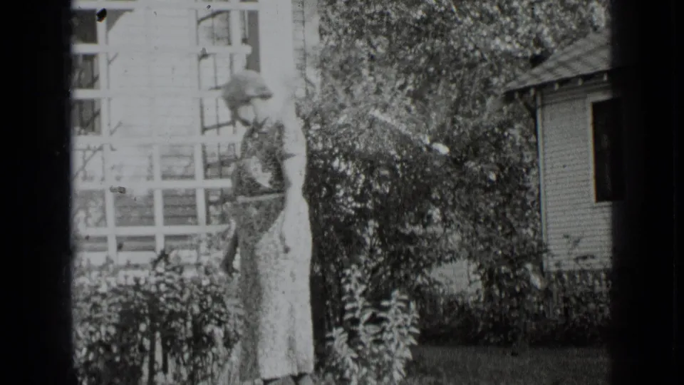 Adult woman in a long patterned dress walking outside a New York residence in 1939 home-movie footage.