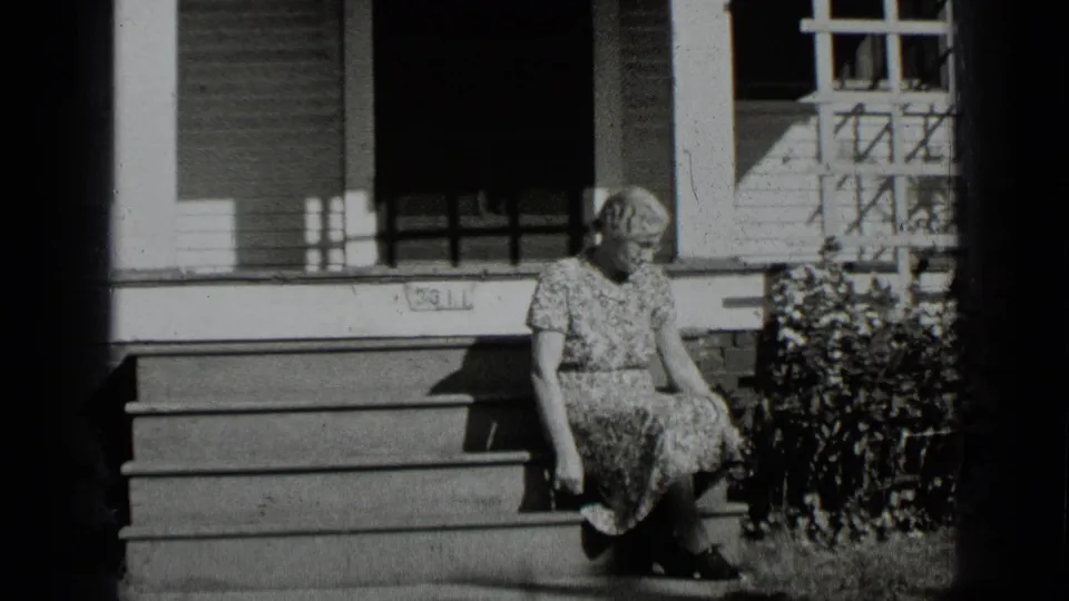Adult woman sits on porch steps outside a New York home, with trellis and siding visible behind her.