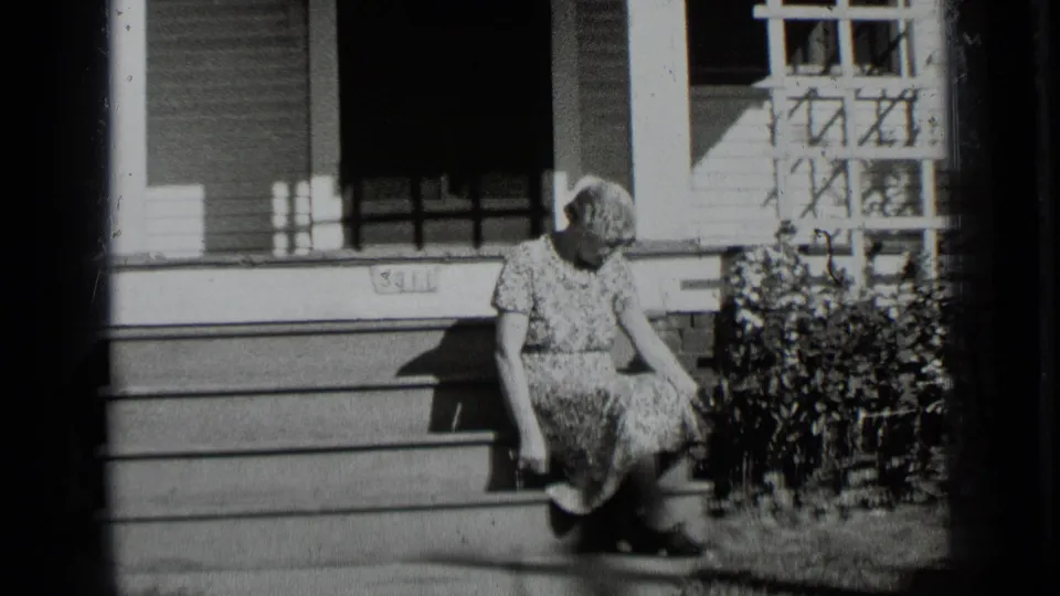 The woman remains seated on the porch steps, head lowered, beside shrubs and a trellis on the house exterior.