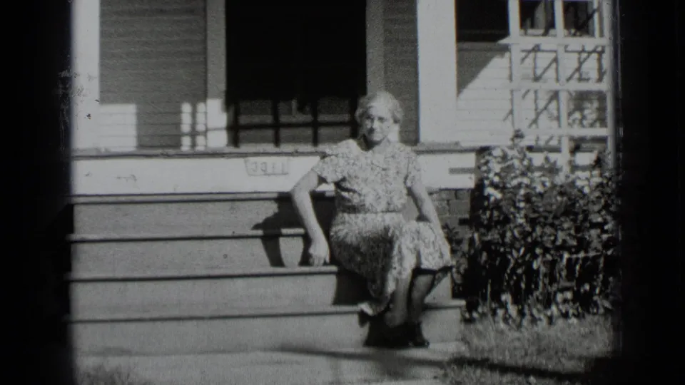 Adult woman in a patterned dress faces the camera while seated on porch steps outside the residence.