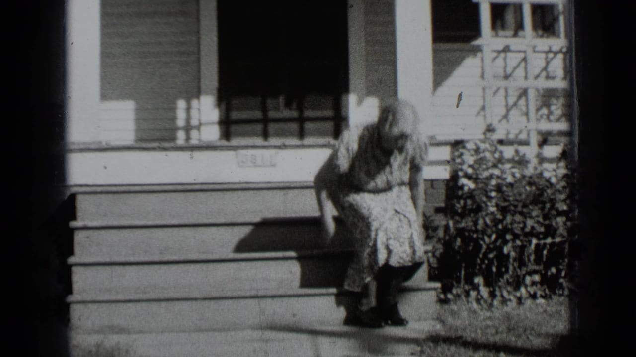 In this 1939 New York home-movie frame, a woman in a floral dress sits on porch steps beside a hedge and shuttered windows.