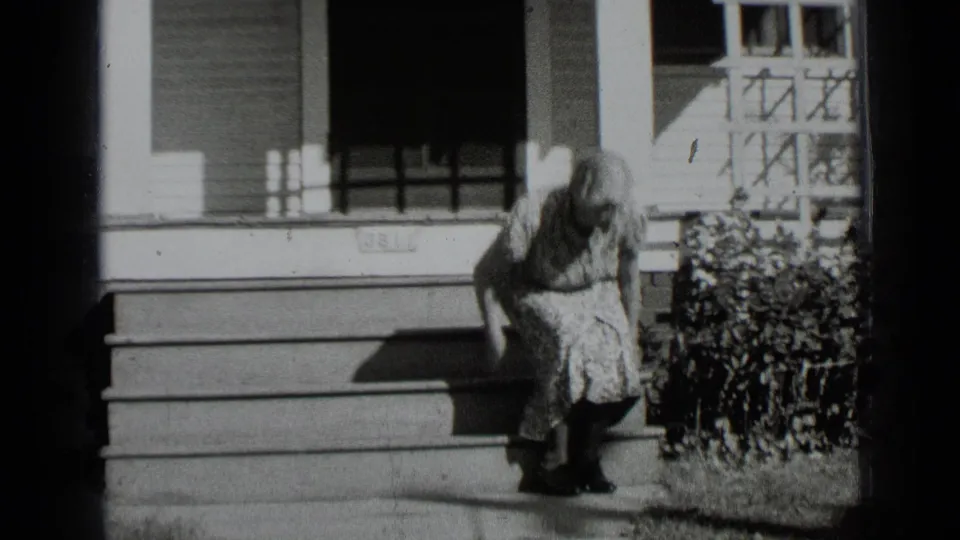 In this 1939 New York home-movie frame, a woman in a floral dress sits on porch steps beside a hedge and shuttered windows.