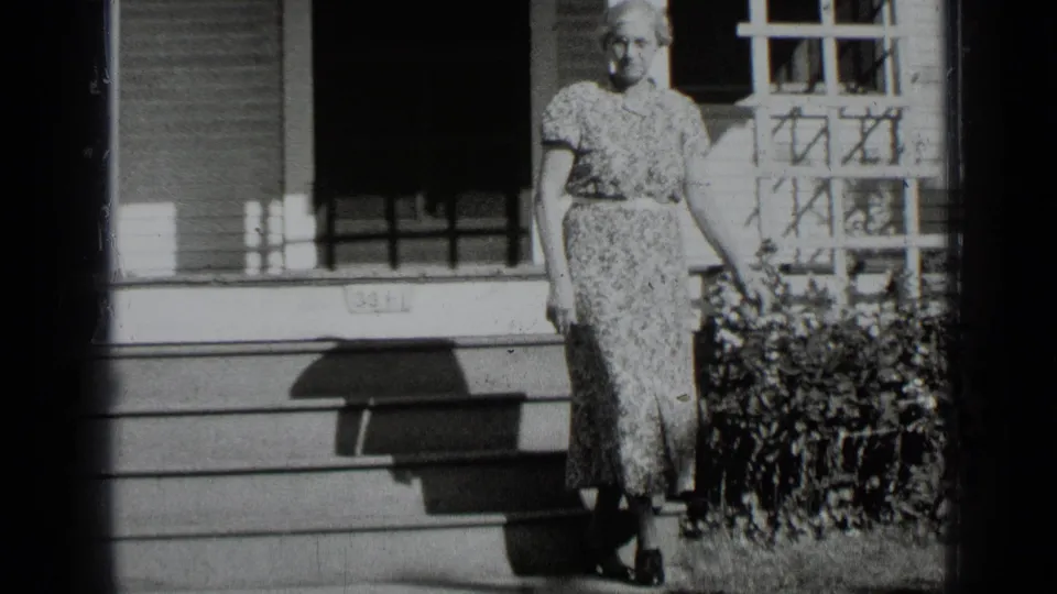 A woman in a floral dress stands near the porch of a New York residence in this supported 1939 black-and-white frame.