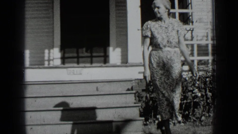 The woman walks down from the porch area, with shuttered windows, siding, and a hedge visible behind her.