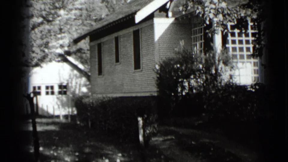1939 New York home-movie frame of a white house exterior with porch glazing, trimmed hedges, trees, and dappled sunlight.