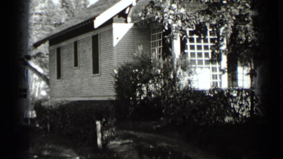 1939 New York 8mm frame with clapboard siding, narrow side windows, bright porch glazing, shrubs, hedges, and a walkway.