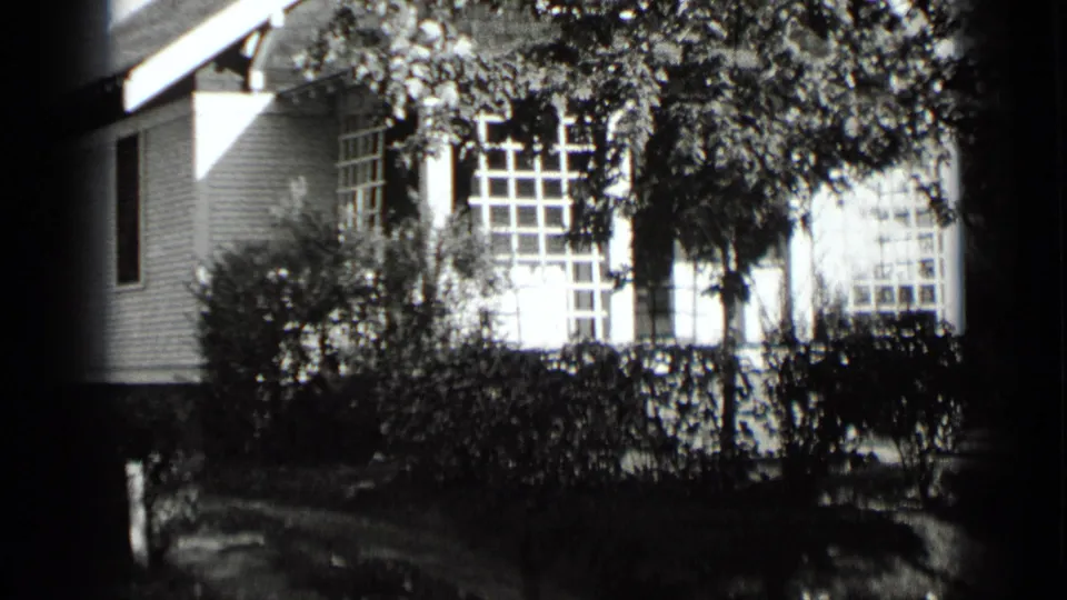 Final 1939 New York frame showing bright porch windows, leafy shrubs, hedges, tree branches, and the shaded front of a white house.