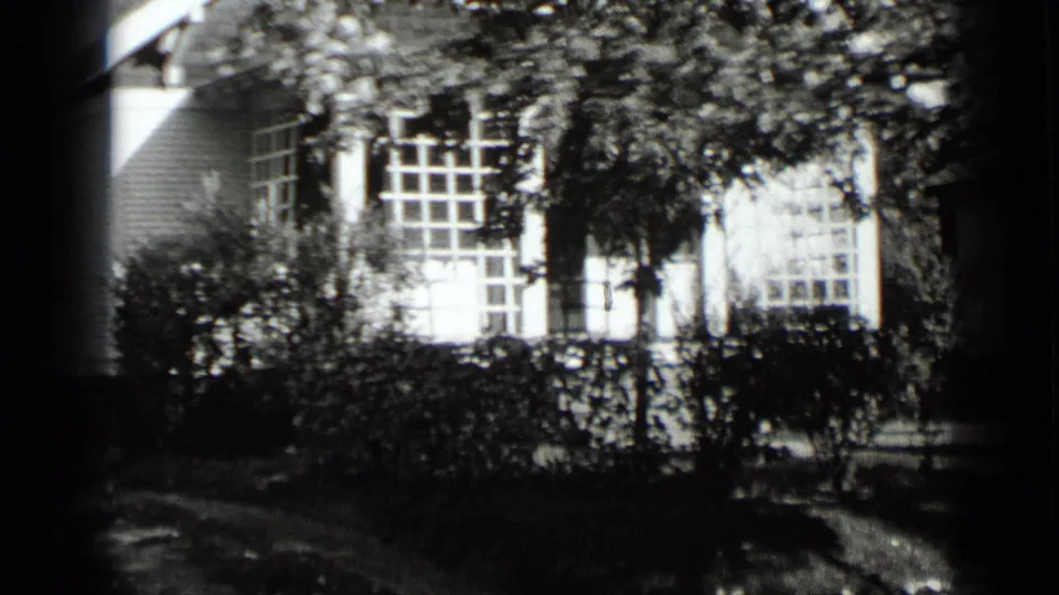 1939 New York home-movie frame showing a suburban house facade with large windows, leafy branches, hedges, and dark foreground framing.