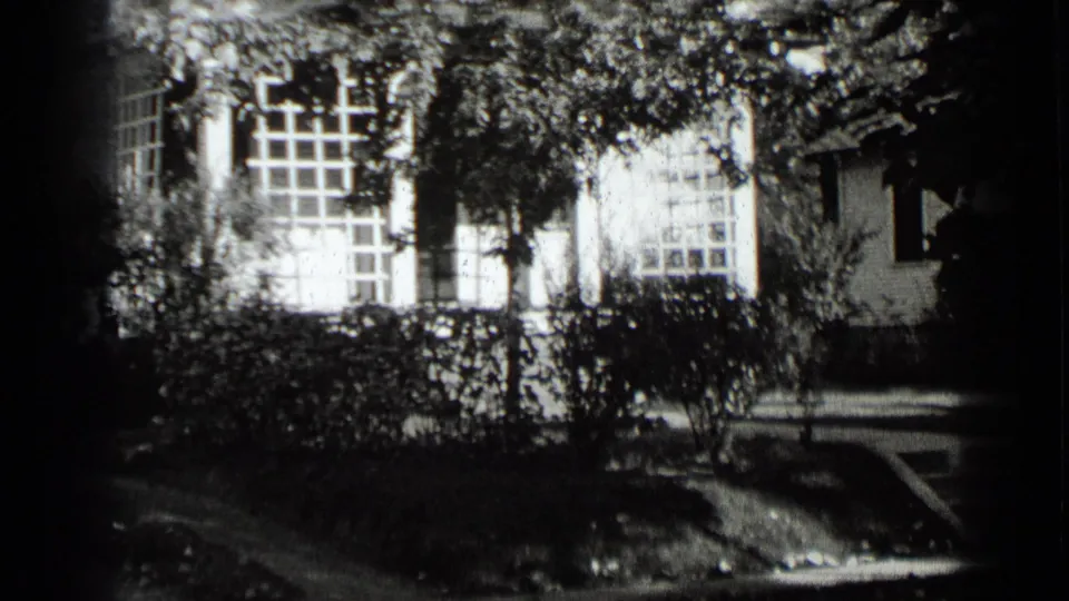 1939 New York suburban street view with a house facade, large windows, leafy trees, trimmed hedges, and a paved walk or drive at the right.