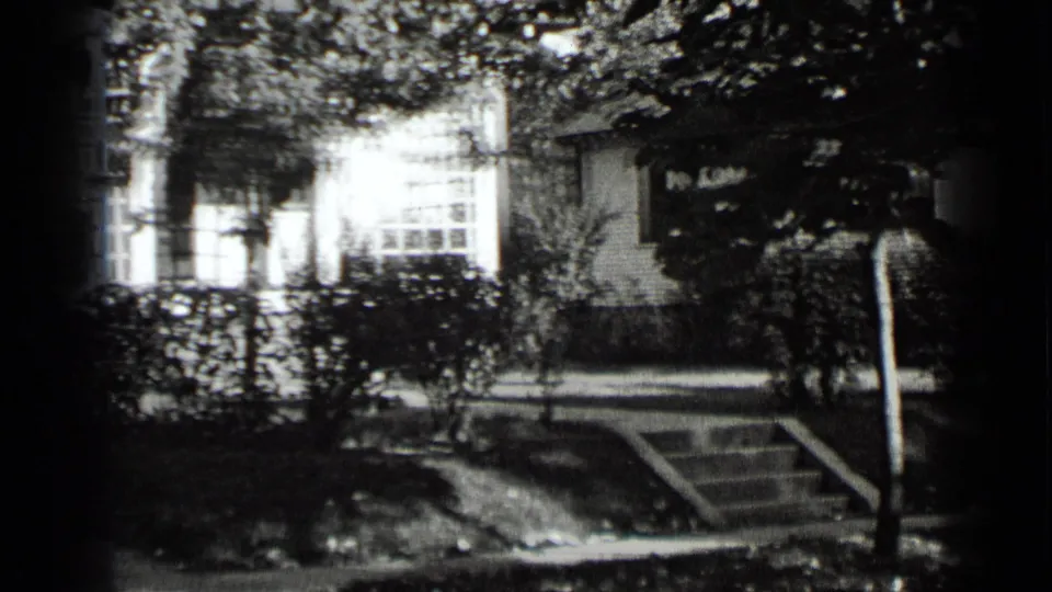 1939 New York home-movie frame showing a suburban house front, hedge line, front steps, tree branches, and a street sign near the curb.