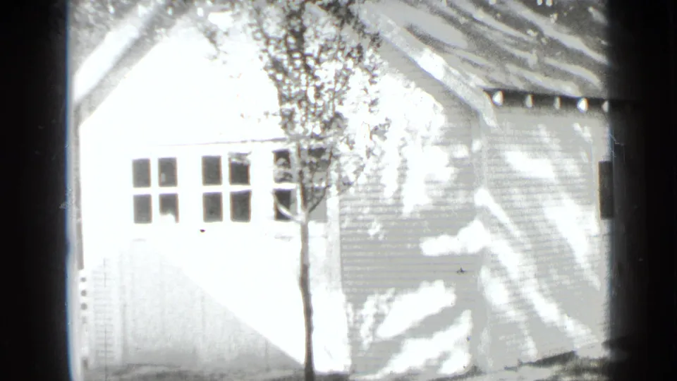 1939 New York residential exterior showing a bright garage or side structure with siding, small windows, a young tree, and hard sun shadows.
