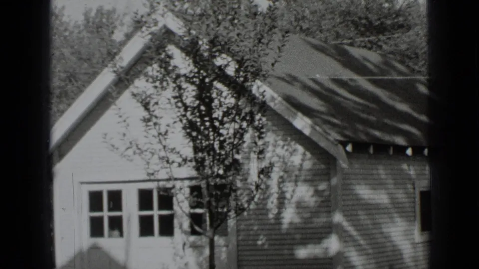 Black-and-white 8mm view of a New York house exterior in 1939, with a tree and shadows across the clapboard wall.