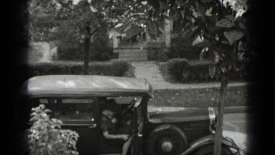 1939 New York home-movie frame with a rounded vintage automobile, two indistinct people, sidewalk, hedges, and nearby houses.