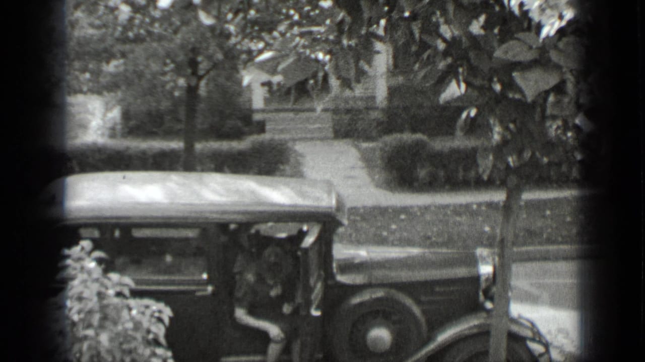 Woman partly visible beside a parked vintage car on a residential street.