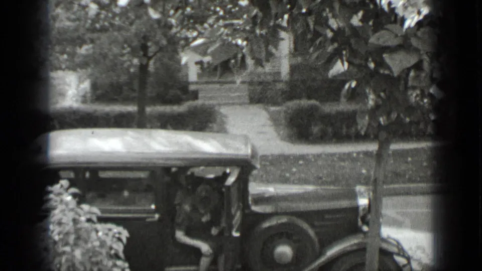 In 1939 New York home-movie footage, a woman stands partly obscured beside a parked vintage car near hedges and a sidewalk.