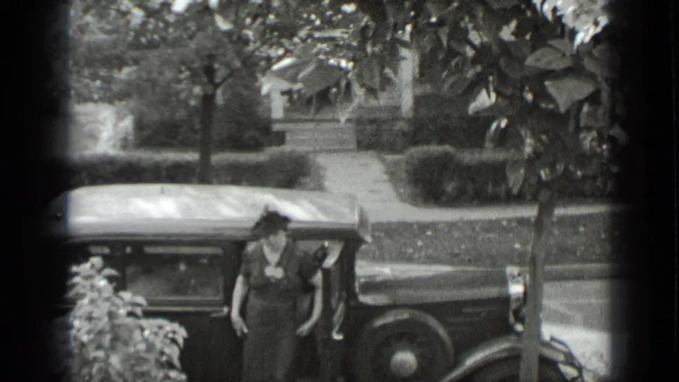 A woman in a dark dress and hat steps from a vintage car on a residential New York street in 1939 footage.