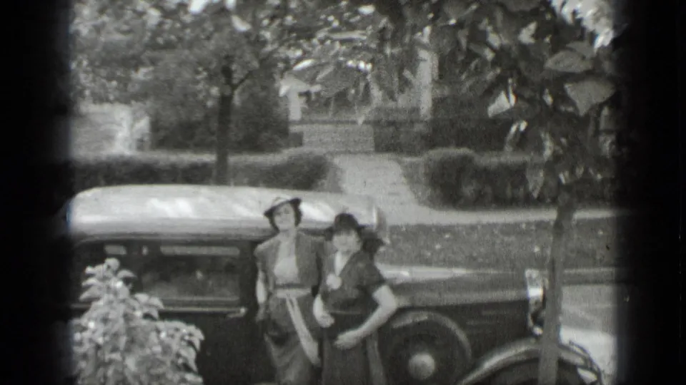 Two women in hats stand near the parked car, with residential lawns, shrubs, and a house visible beyond them.