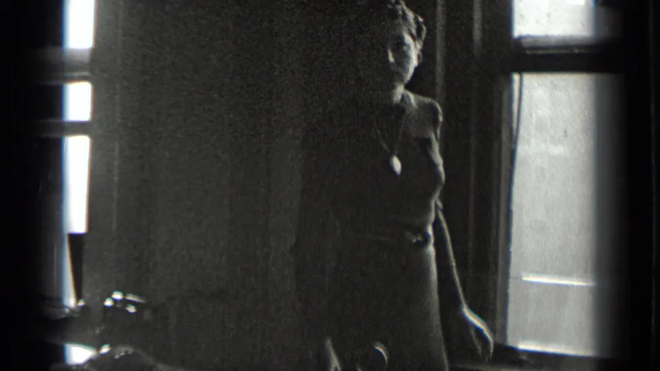 In this 1939 New York home-movie frame, a woman in a dark dress and necklace stands beside a window in a shadowed room.