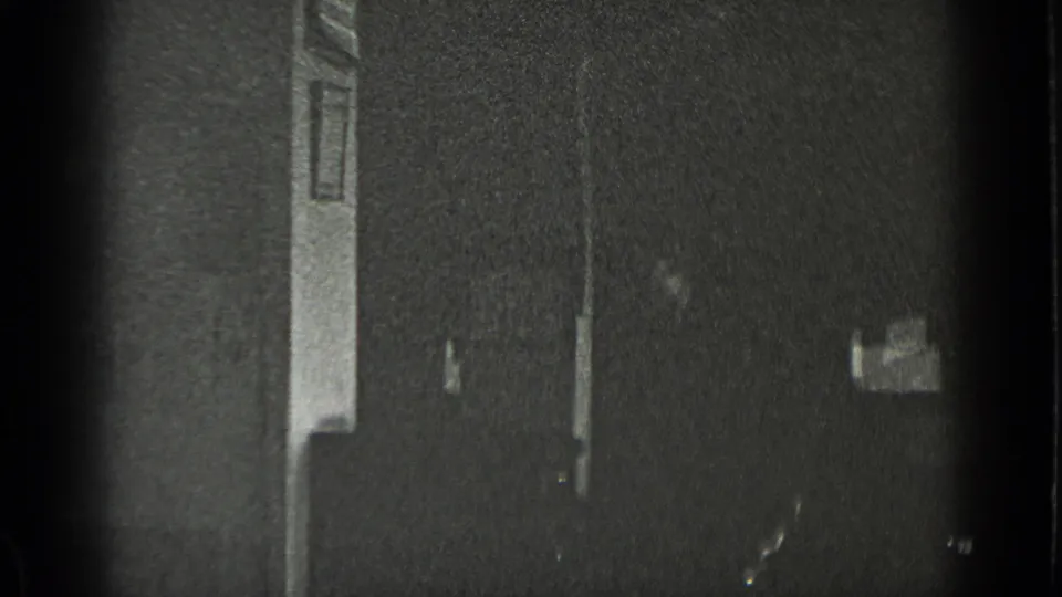 The frame shows a shadowed private interior with doorway trim, a dark wall, and small fixtures visible in soft 8mm focus.