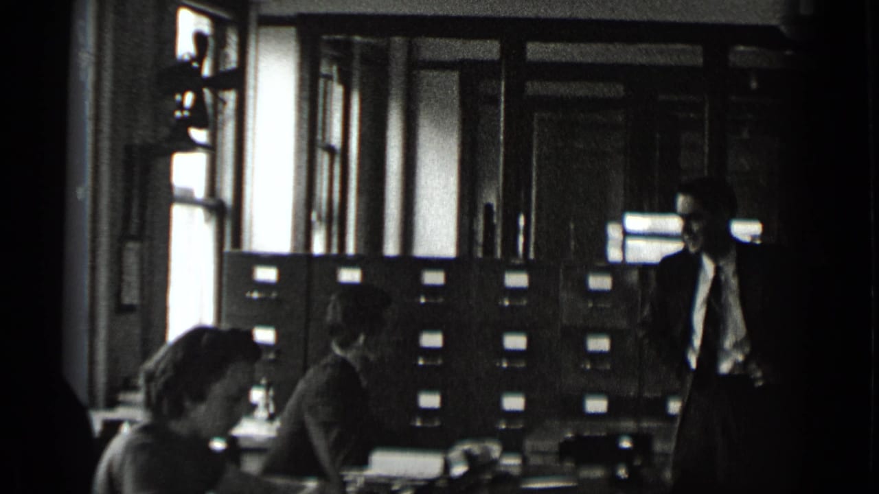 Office workers sit and stand near filing cabinets, desks, and tall windows.